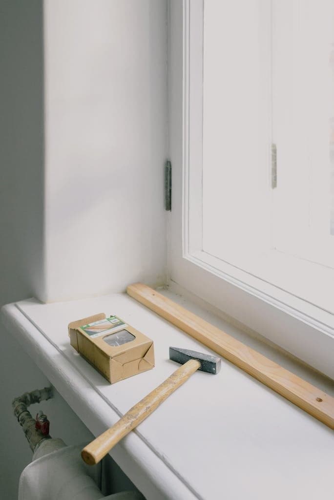 Hammer with wooden handle near plank and cardboard box on windowsill in light house