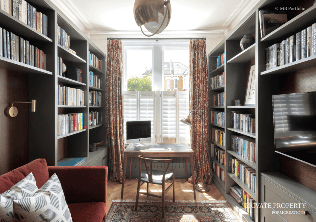 A cozy home office lined with full bookshelves, a desk facing a large window with sheer curtains, a patterned red armchair, and a pendant light, creating a bright, inviting workspace.